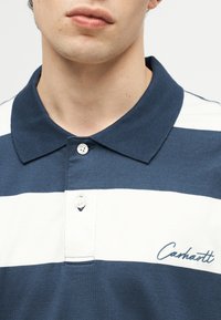 Κοντινό πλάνο ενός ατόμου που φοράει μπλε σκούρο και λευκό ριγέ πόλο με δύο λευκά κουμπιά και κεντημένο λογότυπο "Carhartt".