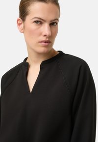 Černý svetr s výstřihem do V. Má raglánové rukávy a hladký povrch. Čisté linie s minimalistickými detaily.