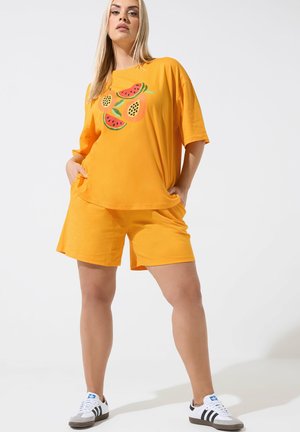 Femme debout les mains dans les poches, portant un short orange vif et un T-shirt assorti avec un motif de fruits, ainsi que des baskets blanches.