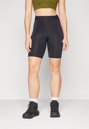 Vrouw draagt zwarte high-waist fietsbroekjes met zijzakken, een olijfgroene crop top, witte sokken en zwarte wandelschoenen.