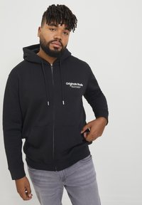 Sort zip-up hoodie med snore og frontlommer. Har hvid "Originals Studio" logo på venstre bryst. Bæres med grå jeans.