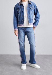 Chaqueta de mezclilla de tela azul claro, cierre de botones, con acentos en rosa en el cuello y los puños. Pantalones vaqueros azul claro con rayas laterales estampadas. Zapatillas blancas.