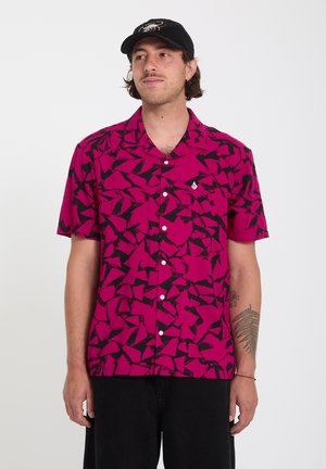 Uomo che indossa una camicia a maniche corte color rosa acceso con motivi astratti neri e un cappello nero, in piedi davanti a uno sfondo semplice.