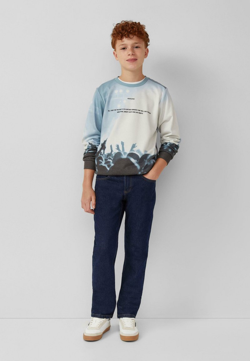 Sudadera gráfica azul claro y blanca con silueta de multitudes, diseño de texto, puños acanalados, combinada con jeans azul oscuro y zapatillas blancas.