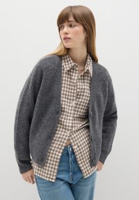 Cárdigan gris de punto con textura acanalada, de corte relajado y con cierre de botones. Llevado sobre una camisa a cuadros en tonos beige y marrón.