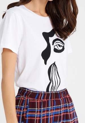 Kvinde iført en hvid t-shirt med abstrakt sort ansigtskunst og en højtaljet plisseret nederdel i rød, blå og sort ternet mønster.