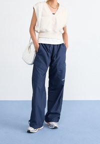 Vit ärmlös topp med V-ringning, marinblå cargobyxor med fickor och silverfärgade träningsskor. Inkluderar en vit väska med kedjerem.