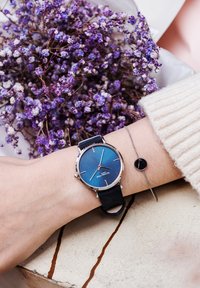 Montre avec un cadran bleu, lunette en argent et bracelet en silicone noir, accompagnée d'un délicat bracelet en argent ornés d'un petit charme circulaire noir.