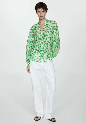 Donna in camicia lunga a maniche verdi floreale, pantaloni bianchi a gamba larga e sandali neri aperti, in piedi contro uno sfondo chiaro uniforme.