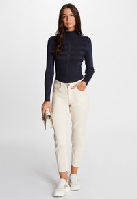 Marineblauwe getailleerde coltrui met gestructureerde strepen, gecombineerd met crèmekleurige high-waisted broek. Model houdt een kleine handtas vast en draagt witte sneakers.