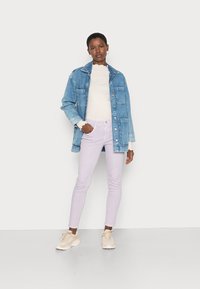 Jaqueta de denim azul claro, suéter branco e calças jeans lilás. Ténis beije. Modelo em pé com as mãos nos quadris contra um fundo simples.