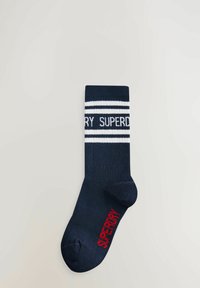 Námořnicky modrá bavlněná ponožka s bílými pruhy a logem "SUPERDRY". Obsahuje žebrovanou strukturu a zaoblený špičku. Akcentní logo v červené barvě na boku.