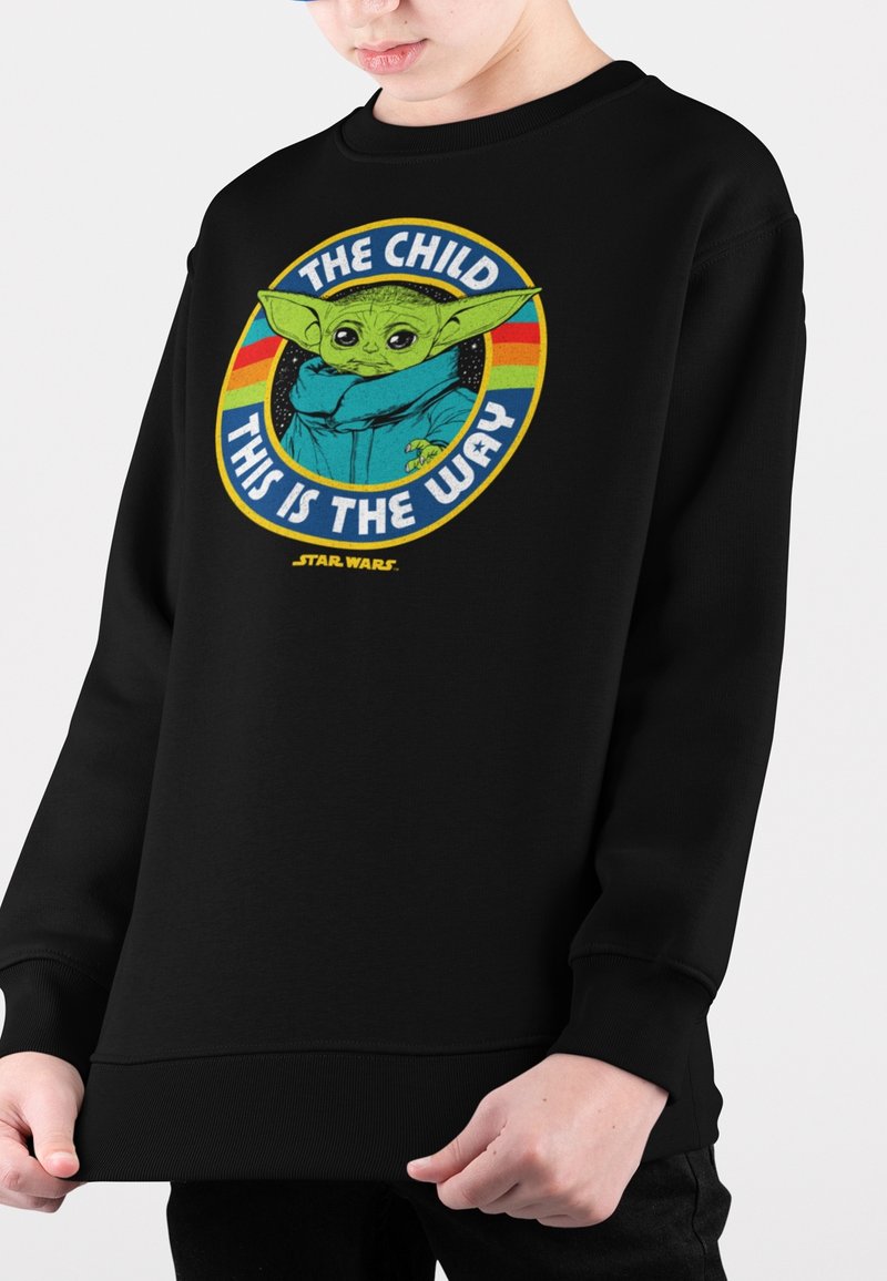 Kind trägt einen schwarzen Pullover mit einem bunten Baby Yoda-Grafik und dem Text "Das Kind, so ist der Weg" aus Star Wars.