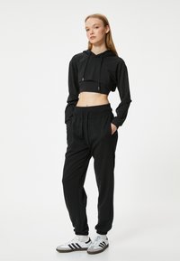 Zwarte cropped hoodie met trekkoord, gecombineerd met zwarte tapse joggers met een elastische tailleband. G gedragen met witte en grijze sneakers.