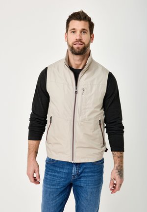 Mann mit Bart und Tattoos, der eine beige ärmellose Weste über einem schwarzen Langarmshirt und blauen Jeans trägt, steht vor einem einfarbigen weißen Hintergrund.