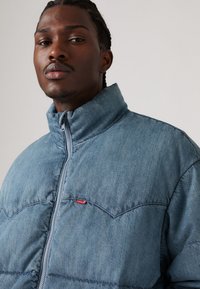 Denim pufferjakke i lysblå, med høj krave, lynlås foran og rød Levi's-logo på brystet, samt en quiltet tekstur.
