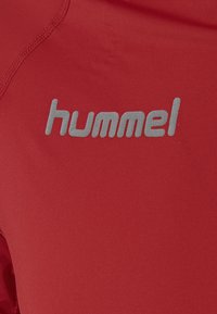 Rode sportshirt van glad materiaal, met een grijs 'hummel' logo op de voorkant en zichtbare stiksels.