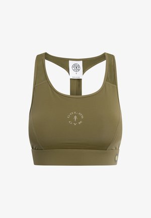 Kobieta stojąca i uśmiechająca się, ubrana w oliwkowozielony sportowy biustonosz i dopasowane legginsy z wysokim stanem oraz białe trampki na białym tle.