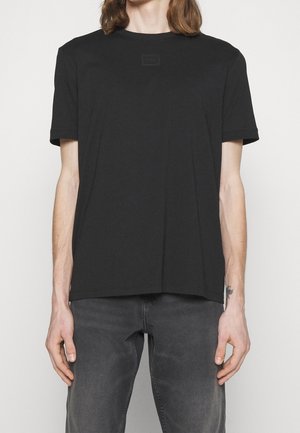Persoon die een effen zwart T-shirt met korte mouwen en donkergrijze spijkerbroek draagt, staand tegen een lichte achtergrond.