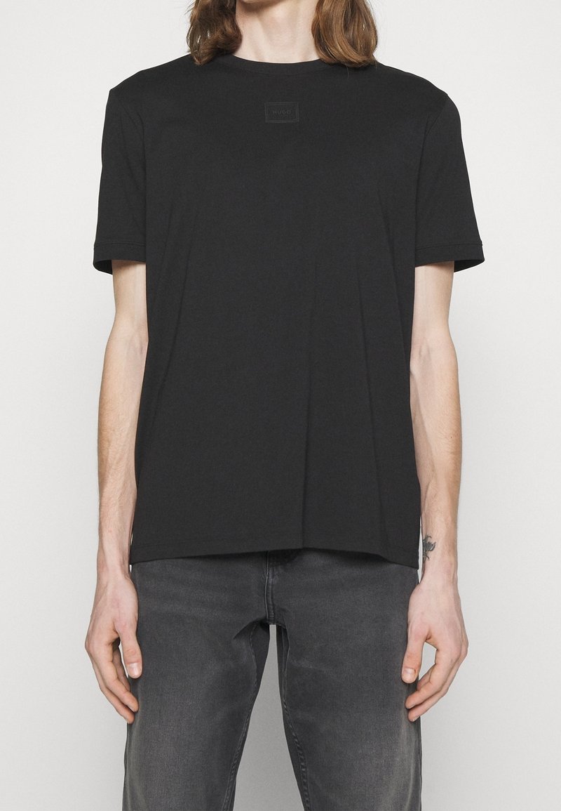 Personne portant un t-shirt noir uni à manches courtes et un jean gris foncé, debout devant un fond clair.