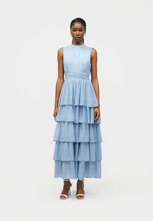 Mujer con cabello trenzado, vestida con un maxi vestido de volantes azul claro sin mangas y tacones altos blancos de punta abierta, de pie contra un fondo liso.