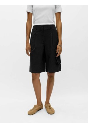 Persoon draagt zwarte cargoshorts tot op de knieën, wit shirt met korte mouwen, beige suède veterschoenen, met ontspannen armen langs het lichaam.