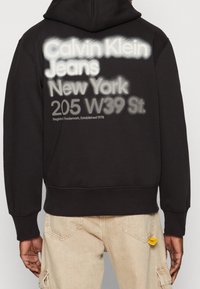Uomo che indossa un hoodie nero con testo bianco e grigio "Calvin Klein Jeans New York 205 W39 St." sulla schiena, abbinato a pantaloni beige.