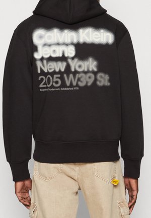 Uomo che indossa un hoodie nero con testo bianco e grigio "Calvin Klein Jeans New York 205 W39 St." sulla schiena, abbinato a pantaloni beige.