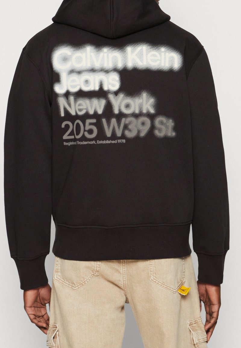 Uomo che indossa un hoodie nero con testo bianco e grigio "Calvin Klein Jeans New York 205 W39 St." sulla schiena, abbinato a pantaloni beige.