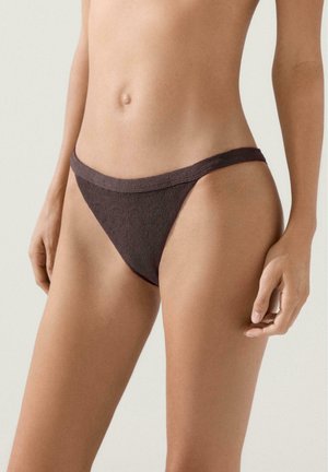 Žena v tmavě hnědých texturovaných spodních dílech bikin, zobrazená od pasu po horní část stehen na jednobarevném světlém pozadí.