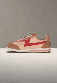 Μπεζ και καφέ αθλητικό παπούτσι με κόκκινο ζιγκ ζαγκ σχέδιο, λευκά κορδόνια και λευκή σόλα, τοποθετημένο σε ανοιχτή επιφάνεια με απλό γκρι φόντο.