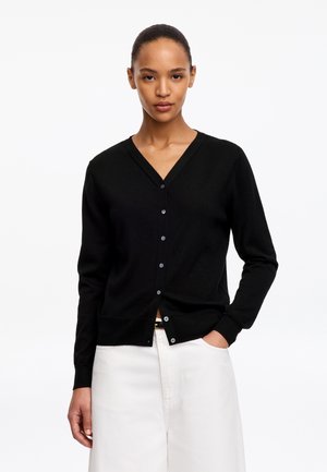 Vrouw draagt een zwarte V-hals knopspeld cardigan en witte broek met één hand in de zak tegen een effen witte achtergrond.
