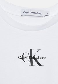 Camiseta blanca de algodón con cuello redondo. Presenta un logo impreso en negro que muestra "CK" y "Calvin Klein Jeans" en diferentes grosores de fuente.
