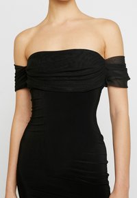 Černé šaty bez ramínek s řasením, přiléhavým střihem a průhlednými texturovanými rukávy, vyrobené ze strečového materiálu.