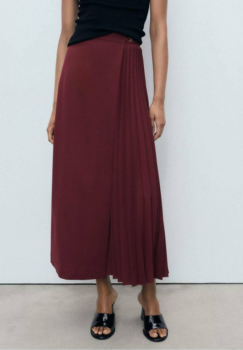 Gonna plissè bordeaux realizzata in tessuto liscio, dotata di chiusura con zip laterale. Abbinata a un top senza maniche nero e sandali neri.