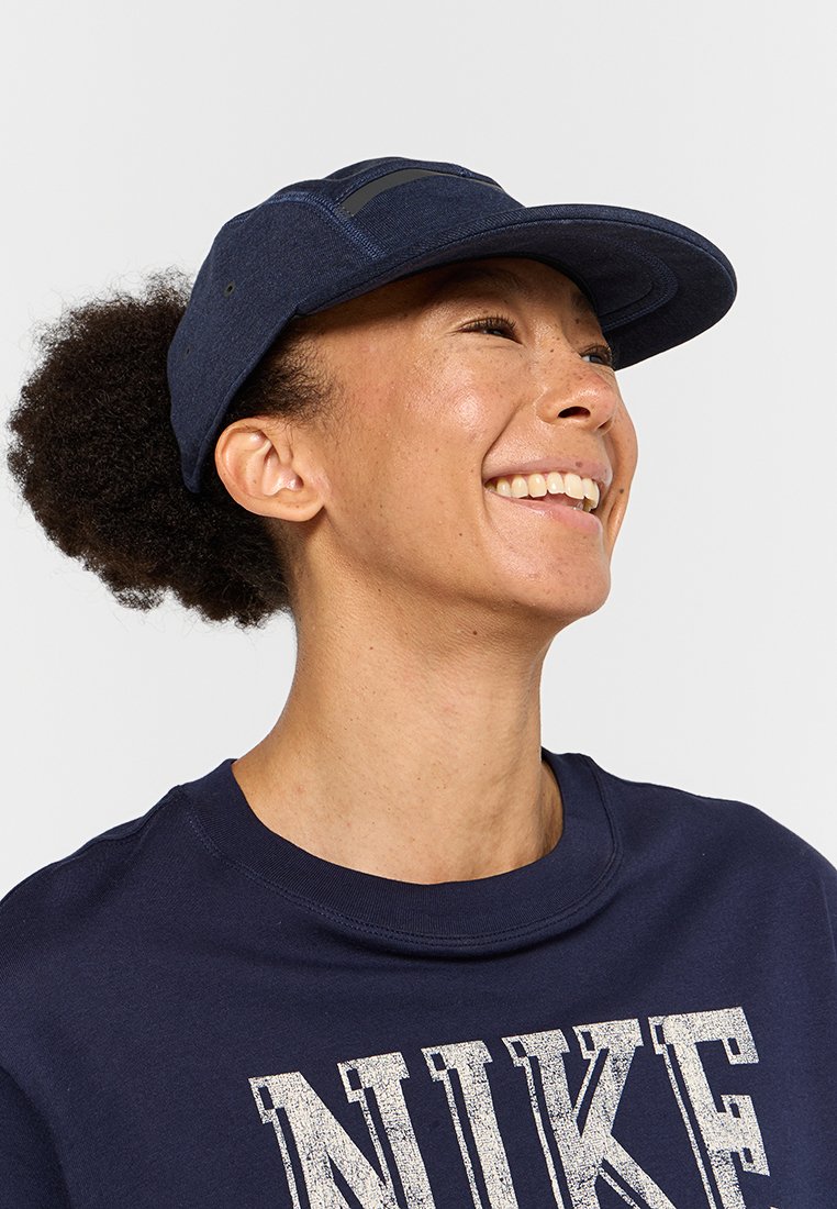Gorra de color azul marino con visera curvada y parte superior plana, que presenta un diseño elegante y sutiles detalles de acento. Se lleva con una sudadera oscura a juego.