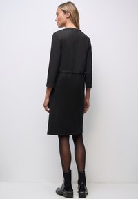 Svart knälång klänning med trekvartslånga ärmar, slät textur, rundad krage och figursydd midja, tillsammans med svarta ankelstövlar.
