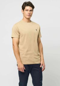 Egy férfi bézs színű, rövid ujjú pólót visel, amelyen egy kis fekete hímzett logó található a mellkasán, sötétkék nadrággal párosítva.