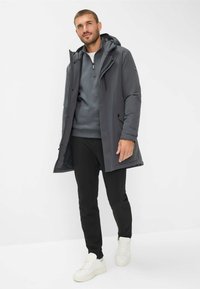 Uomo che indossa un cappotto invernale grigio con cappuccio sopra una felpa grigia a mezza zip, pantaloni neri e sneakers bianche, in piedi su uno sfondo bianco.