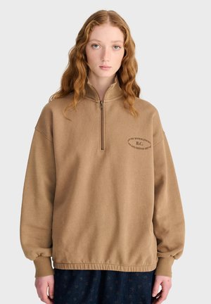 Jeune femme aux longs cheveux roux ondulés, portant un pull marron à fermeture éclair quart de zip, main dans la poche, debout devant un fond clair uni.