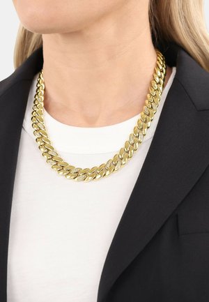 Primer plano de una persona que lleva un collar grueso de cadena dorada con diamantes incrustados, sobre una camiseta blanca y un blazer negro.