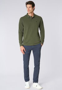 Polo a maniche lunghe verde con colletto, abbinato a pantaloni blu navy e sneakers bianche. Presenta un piccolo logo ricamato sul petto.