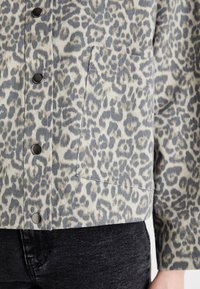 Veste en denim à imprimé léopard avec fermeture à pression, une poche poitrine, et une texture douce dans des tonalités beige, grise et noire.