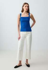 Top a canotta blu, aderente, con scollatura quadrata e tessuto testurizzato; abbinato a pantaloni bianchi a gamba larga e scarpe slip-on nere.