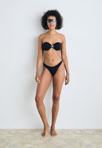 Black bandeau bikini with a front twist detail and metallic accent. Styled with high-waisted bottoms, paired with oversized sunglasses.