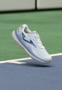 Valge tenniseking siniste aktsentidega, tekstuurse pealisega ja roosa tooni tallaga, esitatud õhus rohelise ja sinise väljakuga taustal.