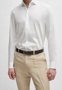 Witte longsleeve met knoopsluiting, gecombineerd met beige broeken en een zwarte leren riem. Het shirt heeft een met patroon gevoerde kraag.