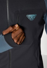 Black athletic jacket featuring a half-zipper, panel design in blue, and a prominent logo on the chest. Includes a zippered pocket.