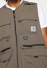 Olivgrön mångficksväst gjord av slitstark tyg, med en framtida dragkedja, olika storlekar av fickor och en märkeslogotyp.