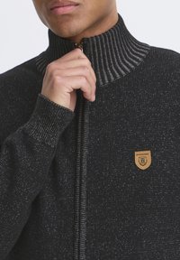 Mustat vetoketjullinen neule takki, jossa on ribbineuleinen kaulus ja hihansuut. Ominaisuuksiin kuuluu teksturoitu neulos ja pieni logopatch vasemmalla puolella.